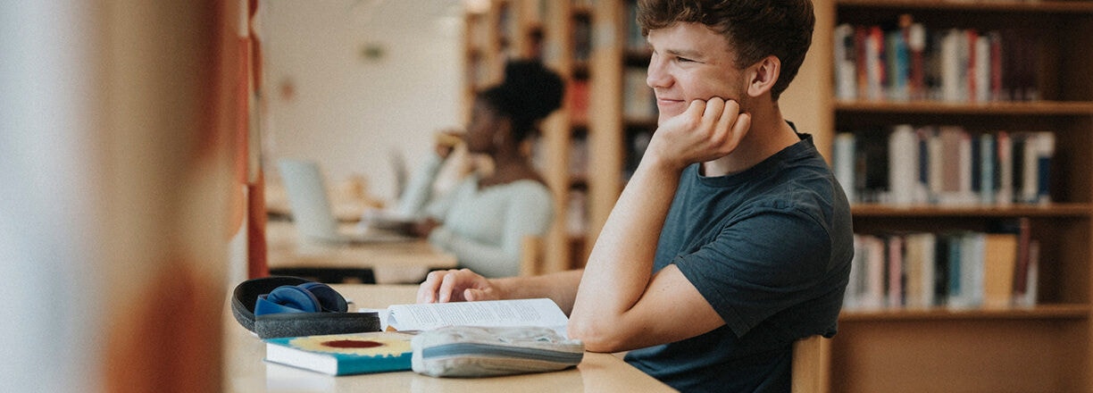 Un estudiante estudiando en un escritorio con libros y un portátil en una biblioteca.