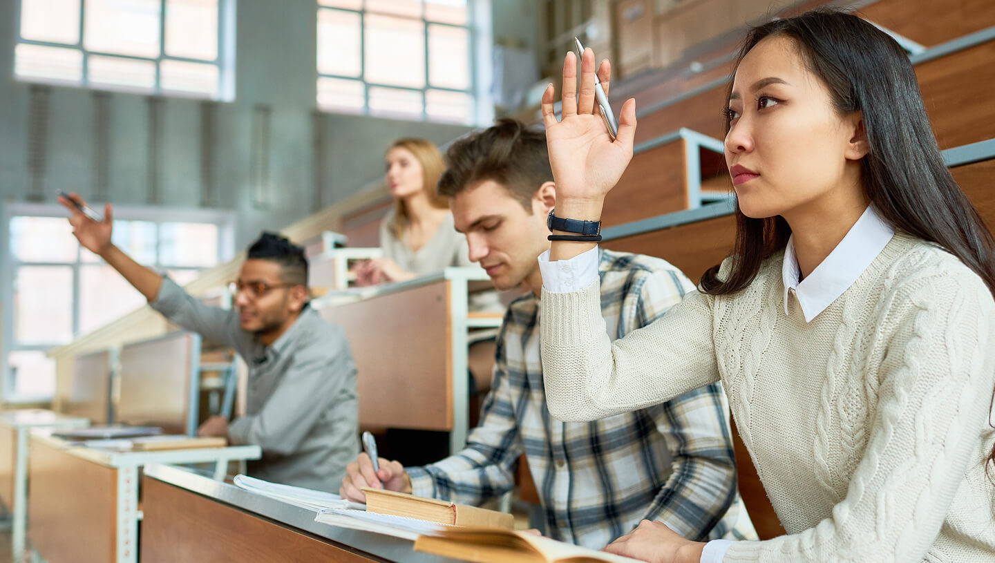 Un aula universitaria con estudiantes sentados en pupitres, mientras una estudiante en primer plano levanta la mano.